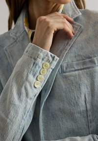 Γυναίκα που διορθώνει το γιακά ενός ανοιχτό μπλε τζιν σακακιού, δείχνοντας τέσσερα κρεμ κουμπιά στο μανίκι και ένα πουκάμισο με σχέδιο από κάτω.