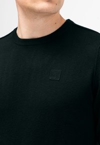 Schwarzer gestrickter Pullover mit Rundhalsausschnitt und kleinem quadratischen Logo auf der Brust. Weiches Gewebe und körpernahe Passform.