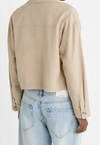 Cropped beige jacket with long sleeves and buttoned cuffs, paired with light blue denim jeans. Smooth, non-textured fabric.
