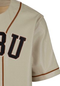Jersey in tessuto beige perforato con lettere ricamate nere "U" e "B" e piping arancione lungo il colletto e i bordi delle maniche.