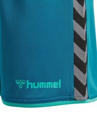 Tyrkysové sportovní šortky s černo-bílými chevron pruhy po stranách a tyrkysovým logem Hummel blízko dolního okraje.