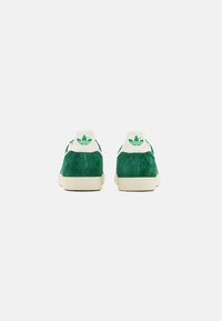 Green suede sneakers with white accents, featuring a logo on the heel and a textured rubber sole, showcasing a low-top design.