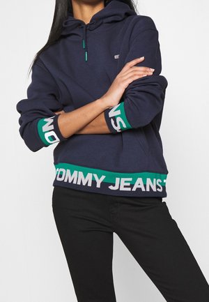 Donna con felpa con cappuccio blu navy con testo verde e bianco "TOMMY JEANS" sui polsini e sull'orlo, abbinata a pantaloni neri, con le braccia incrociate.