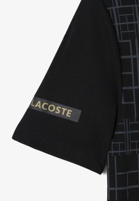 Czarna koszulka z krótkim rękawem w geometryczny wzór. Posiada teksturowaną metkę z logo w złotym kolorze na lewym rękawie. Gładki materiał.