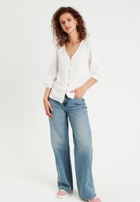 Chemisier blanc à boutons avec un col en V, des manches trois-quarts et un tissu texturé, associé à un jean large bleu clair et des baskets roses.