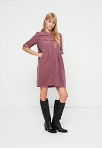 Een knielange jurk met knopen in mauve stof met korte mouwen, voorzien van een geplooide taille en decoratieve stiksels langs de borst.