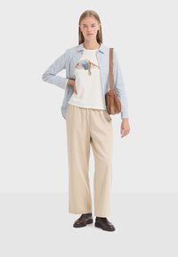White t-shirt with fish graphic, layered under a blue-striped shirt, paired with beige wide-leg pants and brown ankle boots. Crossbody bag accessory.