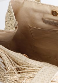 Woven straw handbag with beige fabric interior, featuring a gold snap closure and a side pocket for storage. Fringed detailing on the exterior.