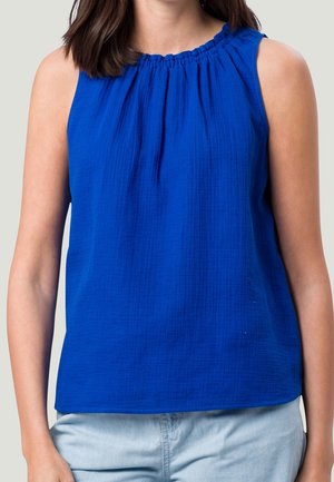 Vrouw draagt een mouwloze, felblauwe top met een gathersluiting bij de hals en lichtblauwe spijkerbroek, staande tegen een effen achtergrond.