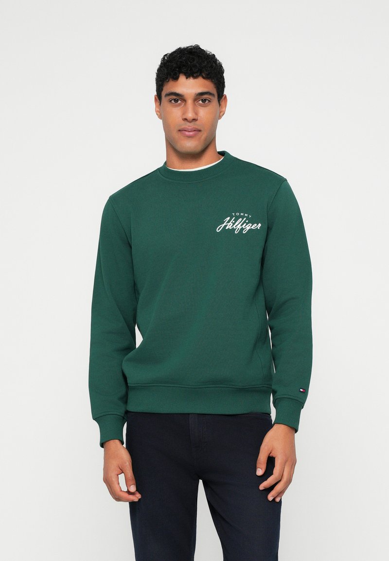 Grønn bomullssweatshirt med lange ermer og rund hals. Har hvit logo-broderi på brystet og ribbestrikkede mansjetter. Kombinert med mørke bukser.