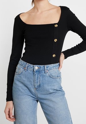 Femme portant un haut noir à manches longues avec trois boutons sur la poitrine et un jean taille haute bleu clair, main sur la hanche.