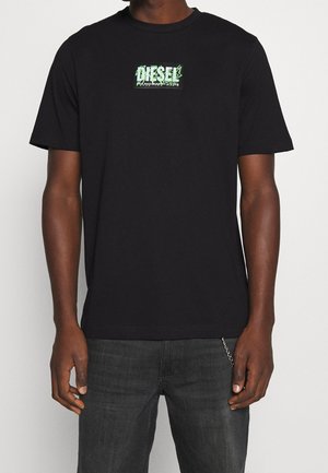 Mand iført sort Diesel t-shirt med neongrønt og hvidt logo centreret på brystet, kombineret med mørkegrå jeans med en kæde som tilbehør.