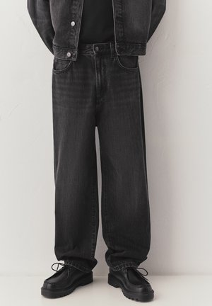 Person iført løstsiddende sorte denimjeans, sort jakke og sorte snørestøvler, stående mod en tekstureret lys væg.