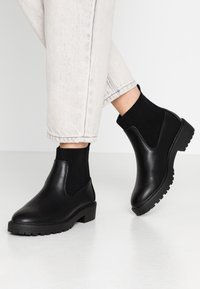 Botas de cuero negro hasta el tobillo con un panel elástico acanalado y una suela texturizada, combinadas con pantalones de mezclilla de color claro, arremangados.