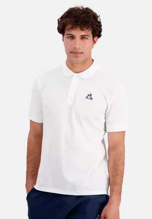 Mand med krøllet hår iført en hvid kortærmet poloskjorte med et lille logo på brystet og marineblå bukser, der står med hænderne i lommerne.