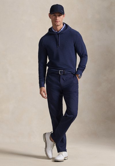 Μπλε ναυτικό φούτερ με κουκούλα και υφή υφάσματος, σε συνδυασμό με μπλε παντελόνι και λευκά παπούτσια. Η εμφάνιση περιλαμβάνει καπέλο και ρολόι στον καρπό.