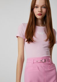 Top rosa de manga corta con textura en canalé, adornado con un diseño de mariposa blanca, combinado con una falda rosa de cintura alta con cinturón y bolsillos traseros.