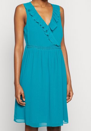 Bezrukankowa turkusowa sukienka z dekoltem w serek, falbanką, haftowanym pasem w talii i marszczonym dołem, noszona przez stojącą osobę na jednolitym tle.