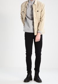 Beige jacka med två bröstfickor ovanpå en grå sweatshirt, i kombination med svarta slim-fit byxor och svarta snörboots. Släta tygtexturer.
