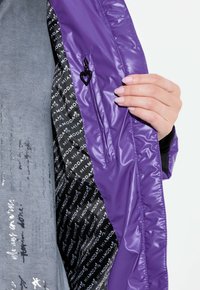 Chaqueta de plumas morada con un acabado brillante, que presenta un pequeño tirador de cremallera en forma de corazón negro y un forro negro con un patrón y texto en blanco.