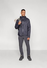 Tamsiai mėlynas vandeniui atsparus striukė su gobtuvu, užtrauktuku ir priekinių kišenių, derinama su pilkais kelnėmis ir juodais žygio batais. Kuprinė per petį.