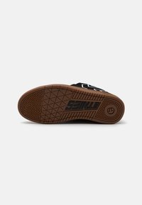 Schwarze Sneakersohle mit einer strukturierten, braunen Gummi-Außensohle, die ein geometrisches Muster und Logo-Details aufweist. Glattes schwarzes Obermaterial oben sichtbar.