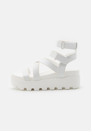 Sandales à plateforme blanches avec larges brides croisées, une bride de cheville ajustable et une semelle texturée pour une meilleure adhérence. Matière lisse avec une finition mate.