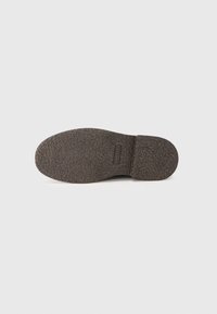 Semelle de chaussure en caoutchouc brun texturé avec un design plat, présentant un bout lisse et une adhérence à motifs pour la traction. Étiquette de marque gravée présente.