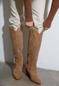 Beżowe zamszowe kozaki do kolan z ozdobnym wzorem motyli i zapięciem na zamek, z blokowym obcasem i spiczastym noskiem.