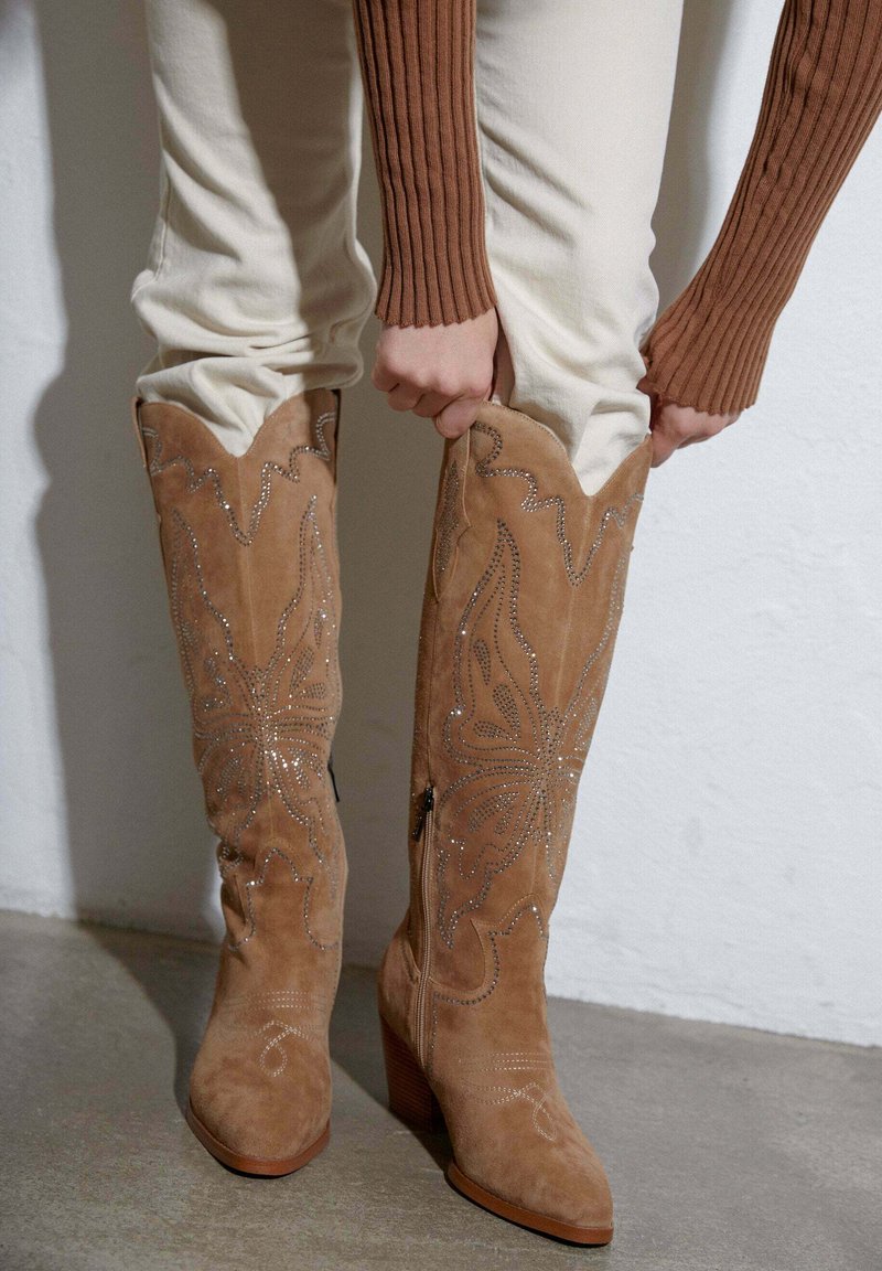 Beżowe zamszowe kozaki do kolan z ozdobnym wzorem motyli i zapięciem na zamek, z blokowym obcasem i spiczastym noskiem.