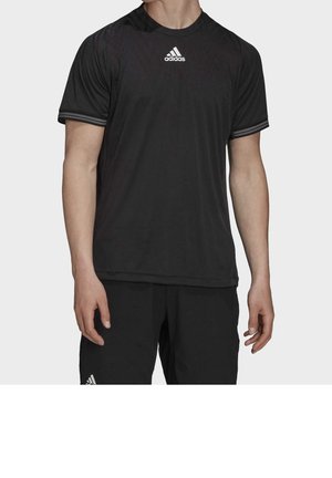 Homme portant un t-shirt Adidas noir à manches courtes et un short noir avec le logo Adidas sur la poitrine et la cuisse gauche sur un fond uni.