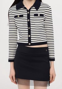 Striped cropped cardigan in navy and cream, featuring buttoned front, two chest pockets, and fitted black skirt with sheer hem.
