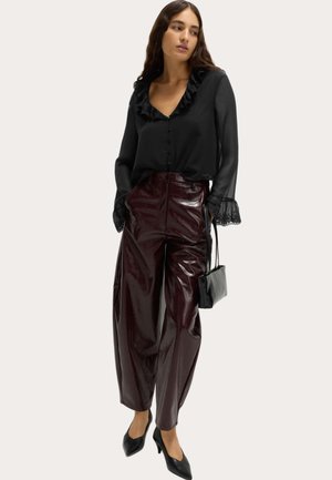 Zwarte blouse met een gerimpelde halslijn en manchetten, gecombineerd met hooggeknoopte, glanzende bordeauxrode broeken. Het model houdt een kleine zwarte handtas vast.