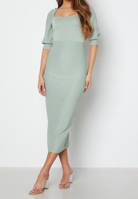 Robe midi vert menthe avec une encolure carrée, des manches bouffantes, une texture côtelée et une silhouette ajustée. Sandales à talons blocs transparents.