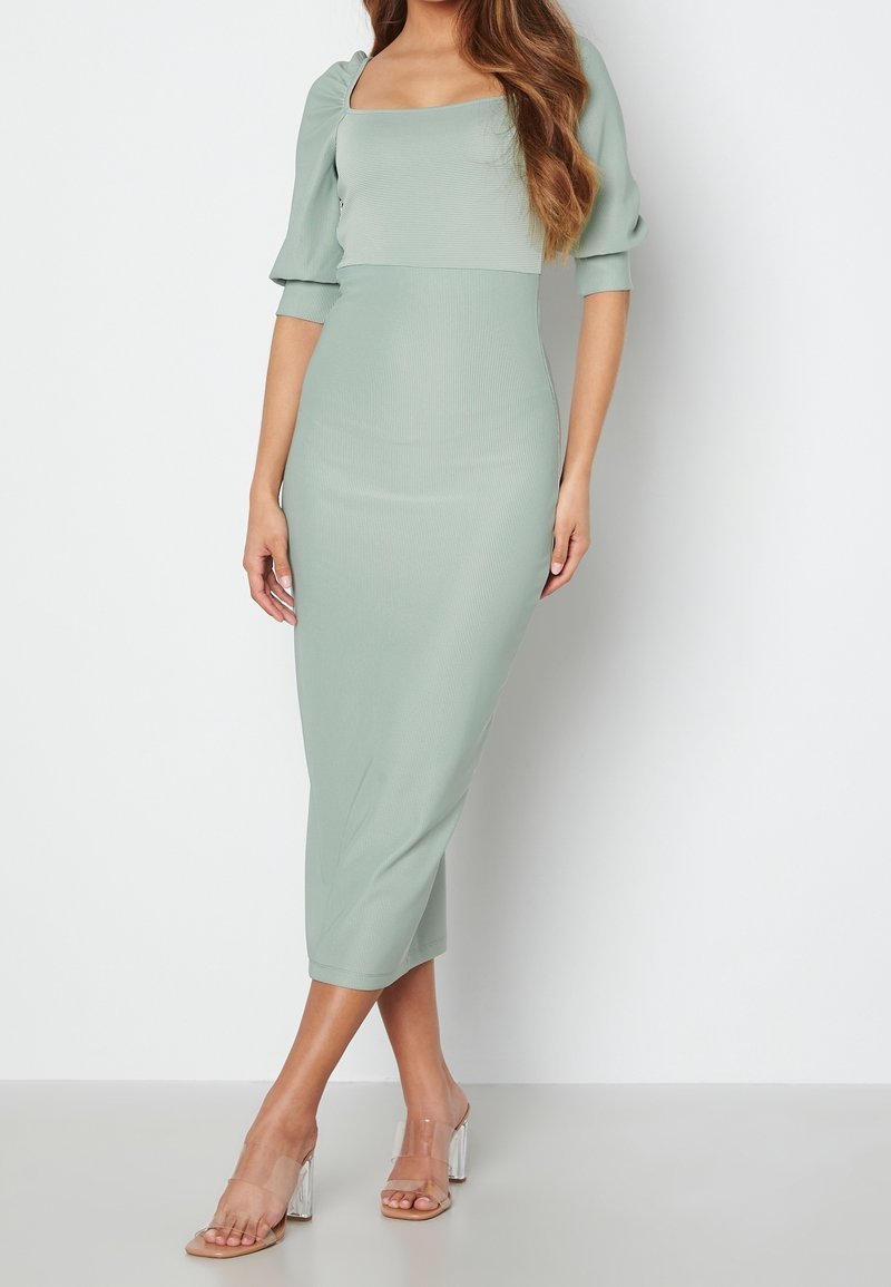 Robe midi vert menthe avec une encolure carrée, des manches bouffantes, une texture côtelée et une silhouette ajustée. Sandales à talons blocs transparents.