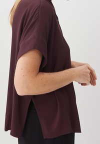 Blusa bordeaux con maniche corte, tessuto morbido, vestibilità ampia, spacchi laterali e scollatura rotonda, indossata su pantaloni neri.