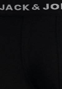 Sous-vêtements noirs en tissu doux avec une texture lisse, mettant en valeur une ceinture contrastante blanche avec logo. Design simple et épuré.