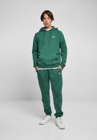 Felpa verde con cappuccio regolabile, tasca frontale e logo. Abbinata a pantaloni jogger verdi, entrambi in tessuto morbido e vestibilità rilassata. Sneakers bianche.
