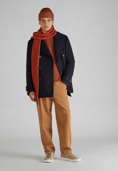 Giacca peacoat marine su un maglione ruggine, abbinata a pantaloni beige e sneakers color crema. Gli accessori includono una sciarpa a coste ruggine e un berretto coordinato.