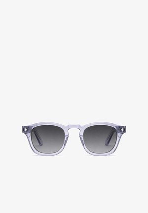 Junger Mann mit Sonnenbrille, rückwärts getragener schwarzer Kappe, schwarz-weiß geblümtem Hemd, steht mit verschränkten Armen vor einfarbigem Hintergrund.