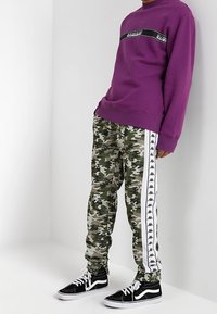 Paarse sweatshirt met zwarte accenten en logo, gecombineerd met groene camouflagebroek met witte zijkanten. Zwarte sneakers maken de look compleet.