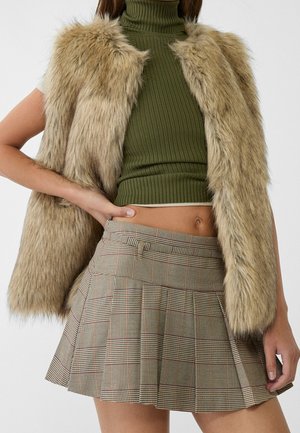 Woman wearing a sleeveless beige faux fur vest, olive green ribbed turtleneck crop top, and pleated plaid mini skirt.