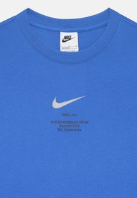 Sinine puuvillane T-särk ümmarguse kaelusega. Omab valget Nike swoosh logo ja musta trükitud teksti, sealhulgas brändi ja aadressi üksikasju.