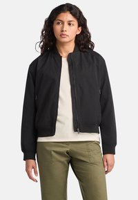 Black bomber jacket with a zip front, ribbed cuffs, and two side pockets. Worn over a light-colored shirt and paired with olive green pants.