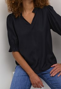 Blouse noire à manches courtes avec un col en V et des accents plissés au niveau de l'encolure. Tissu lisse ; coupe décontractée. Portée avec un jean bleu.
