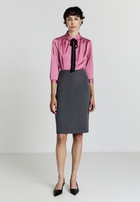 Blouse en soie rose avec un nœud noir, et une jupe crayon ajustée en gris avec des rayures. Des chaussures noires à bout pointu complètent le look.