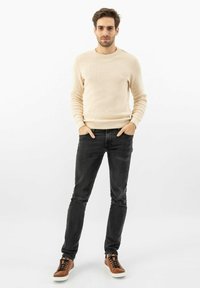 Beige pruhovaný pletený svetr, černé úzké džíny a hnědé tenisky s bílou podrážkou. Modelka stojí se založenýma rukama v kapsách na bílém pozadí.