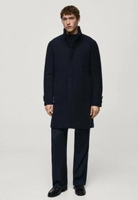 Navy ullrock med hög krage, framtida knäppning och sidofickor. Kombinerad med matchande eleganta byxor och svarta skor.