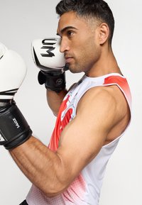 Boxeur portant un débardeur blanc et rouge, gants de boxe noirs, se concentrant sur sa technique. Bras musclés, design spécifique au sport. Détails en tissu texturé visibles.