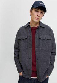 Chaqueta de camisa de lana gris con dos bolsillos en el pecho, cierre de botones y un ajuste relajado sobre un suéter burdeos, combinada con una gorra azul marino.